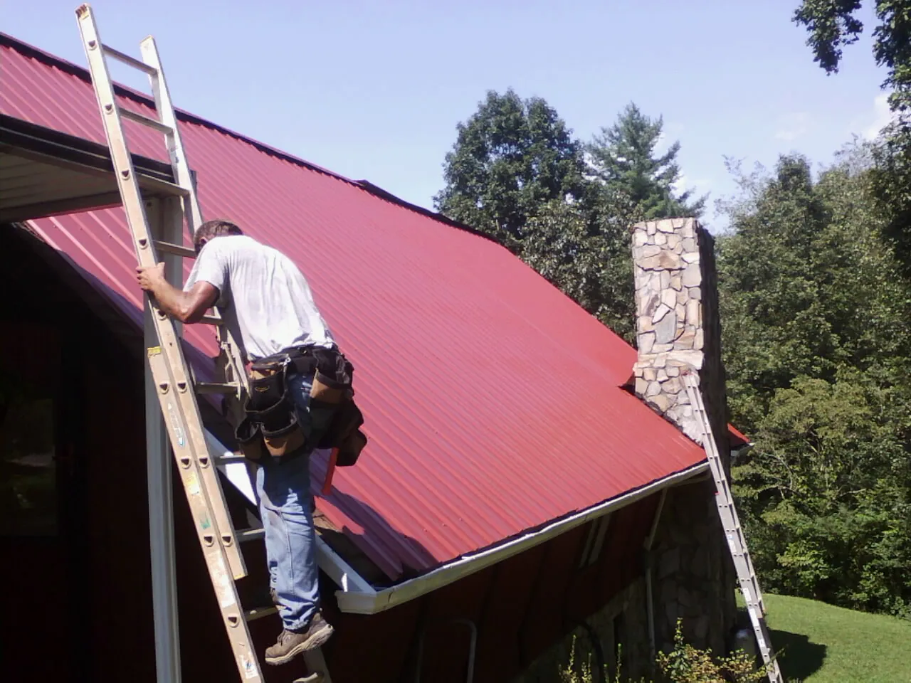 Roofer on a ladder beside a red metal roof and stone chimney — Soffit & Fascia Repair by Roofing Brothers in Pigeon Forge